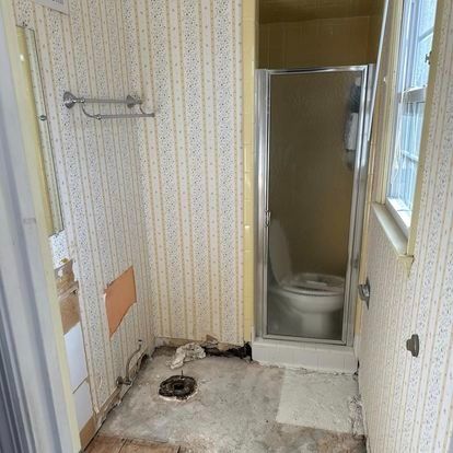 Bathroom undergoing renovation; exposed floor, toilet, shower, and wallpaper visible.