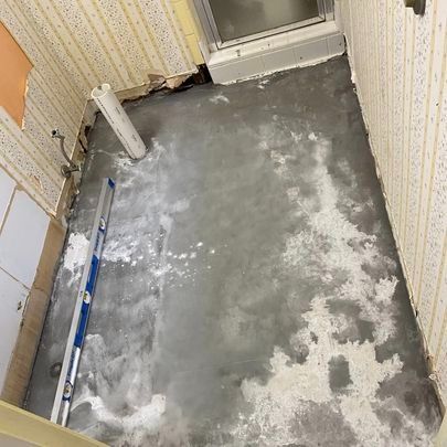 A room with a concrete floor covered in white patches; the walls have patterned wallpaper and exposed wires.
