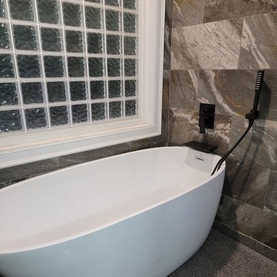 White oval bathtub next to a glass block window and dark gray tile wall. Black faucet and hand shower.