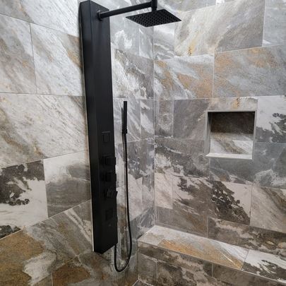 Black shower panel with showerhead and handheld sprayer, mounted on marbled tile wall with recessed shelf.