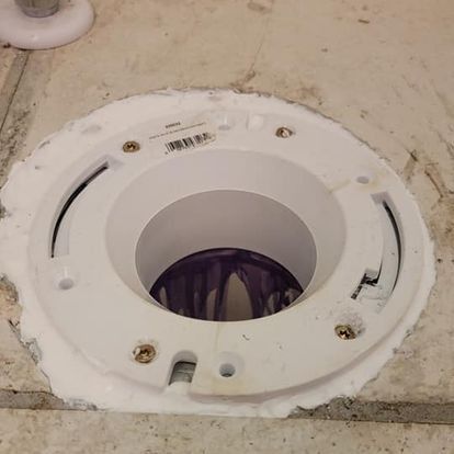 White toilet flange installed on a bathroom floor, secured with screws and sealant.