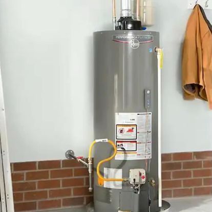 A tall, gray water heater sits against a brick wall in a room, with yellow gas lines connected.