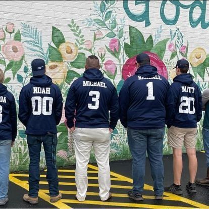 Group of people in navy hoodies with names and numbers on the back, standing in front of a mural of flowers.