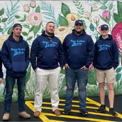 Five men wearing navy hoodies and hats, standing in front of a mural, smiling.