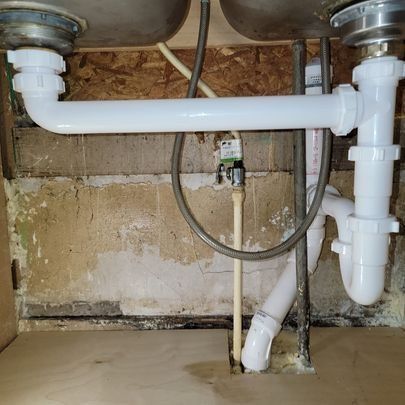 White plumbing pipes and sink drain under a sink, against a weathered wall.