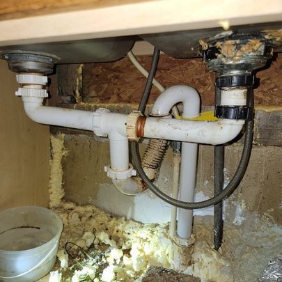 Under-sink plumbing: White pipes connect two basins to drain pipe. Brown cabinet and spray foam visible.