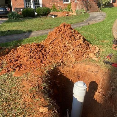 Hole dug in yard with a white pipe. A pile of reddish-brown dirt is next to the hole. Grass and house in background.