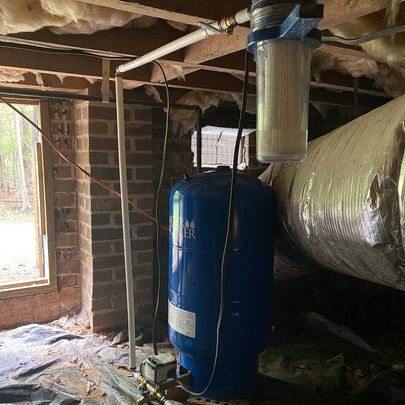 Blue water tank, filter, and plumbing in a crawl space with exposed brick and insulation.