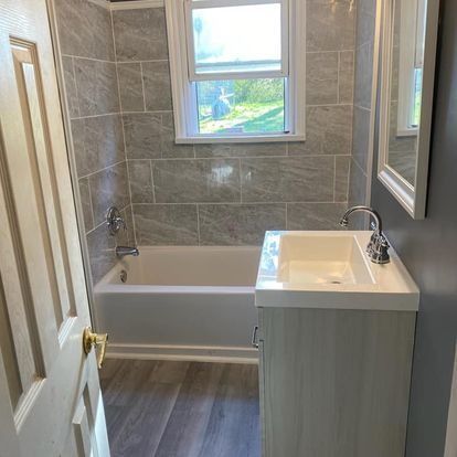 Bathroom with gray tiled walls, a white sink and vanity, and a bathtub under a window.