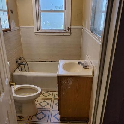 Small, old bathroom with toilet, sink, tub, and window. Walls are cream and white. Floor is patterned.