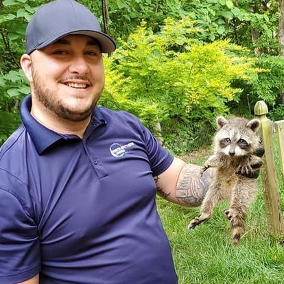 Man in blue shirt and cap holding a raccoon outdoors, smiling.