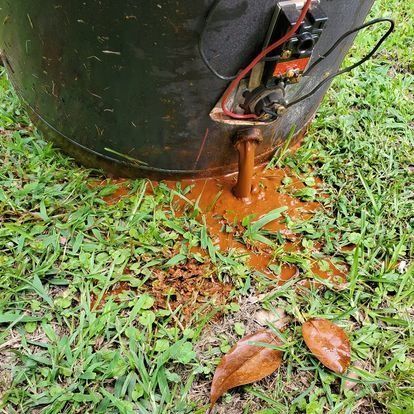 Rusty water leaking from a damaged black water heater on a grassy lawn. Wires exposed.