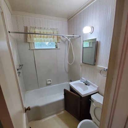 Bathroom with a white tub and sink, toilet, brown vanity, and a small window with a curtain.