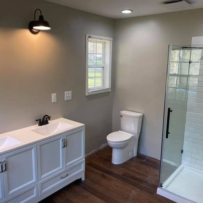 Newly renovated bathroom with a white double vanity, toilet, and glass shower. Gray walls and wood floor.