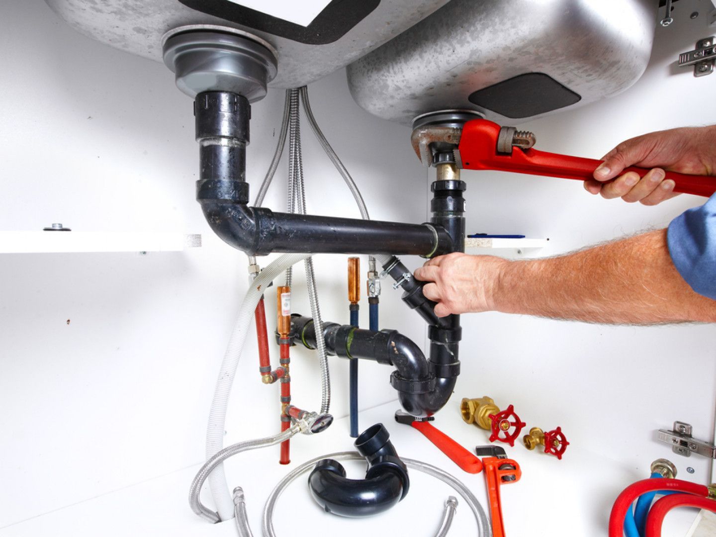Plumber fixing pipes under a kitchen sink with a wrench. White cabinet, silver sink, black and copper pipes.