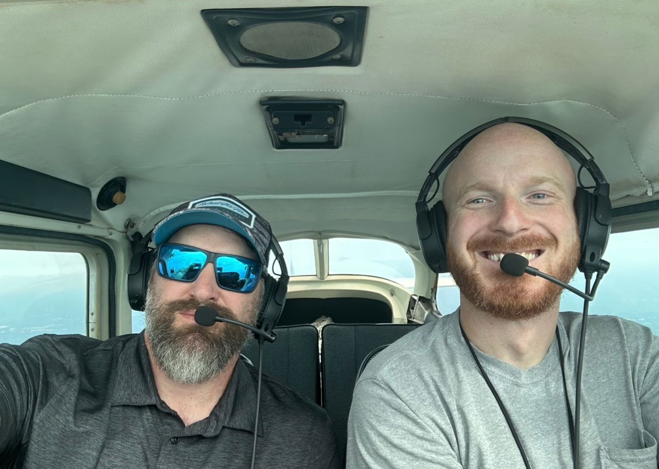Two men are sitting in a plane wearing headphones and sunglasses.