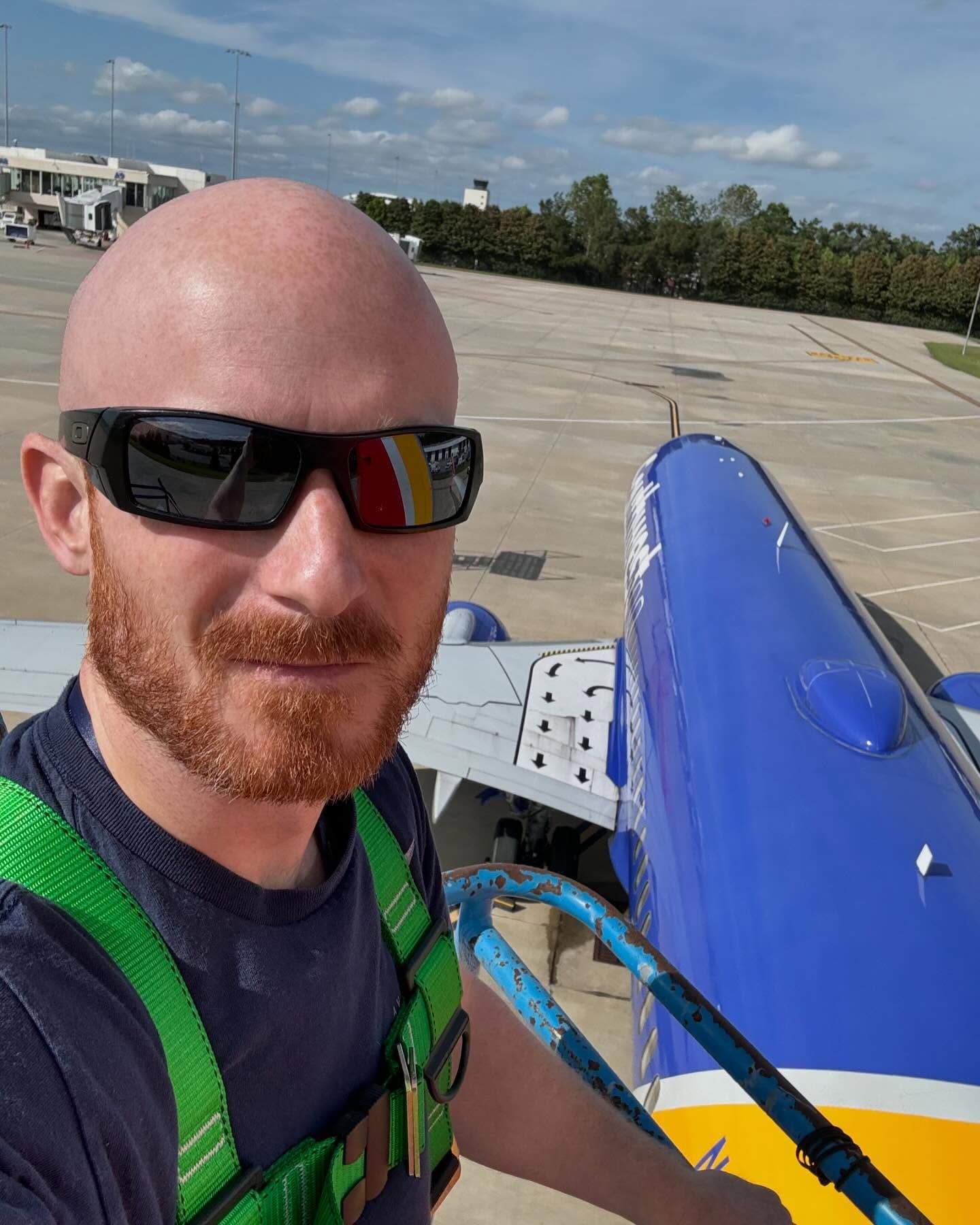 A man wearing sunglasses and a green vest is standing in front of an airplane.