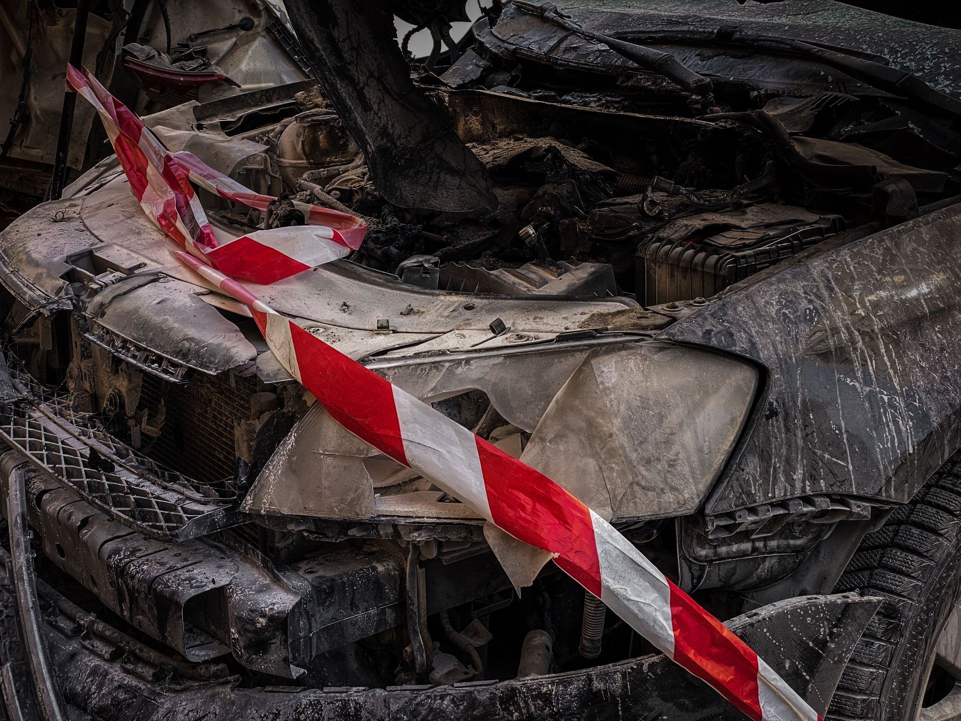 Burned-out car wreck behind red-and-white caution tape, with charred, twisted metal.