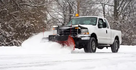 White pickup truck plowing snow on a road with snowy trees in the background.