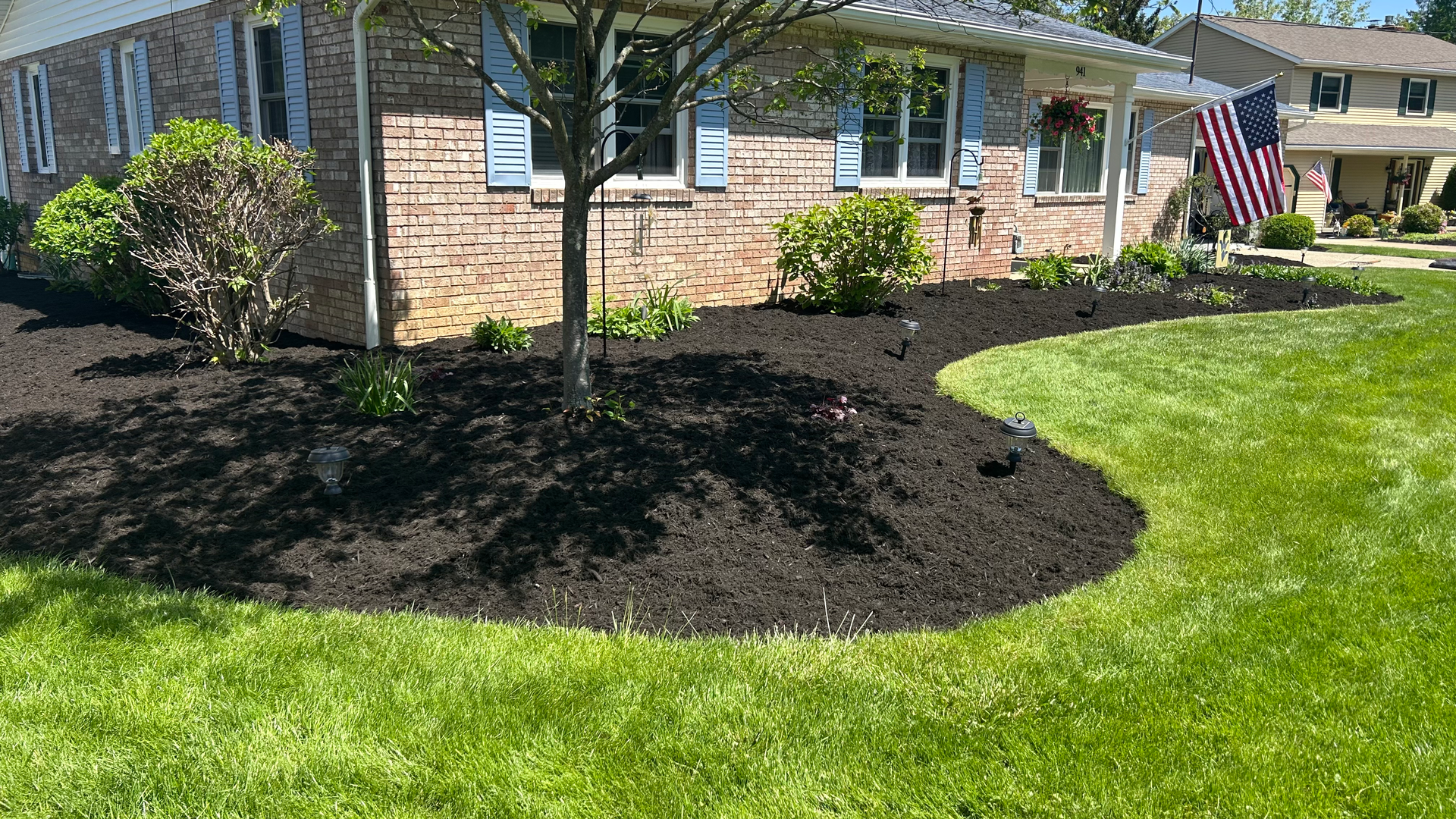 Front yard with a brick house, black mulch flower bed, green grass, and an American flag.
