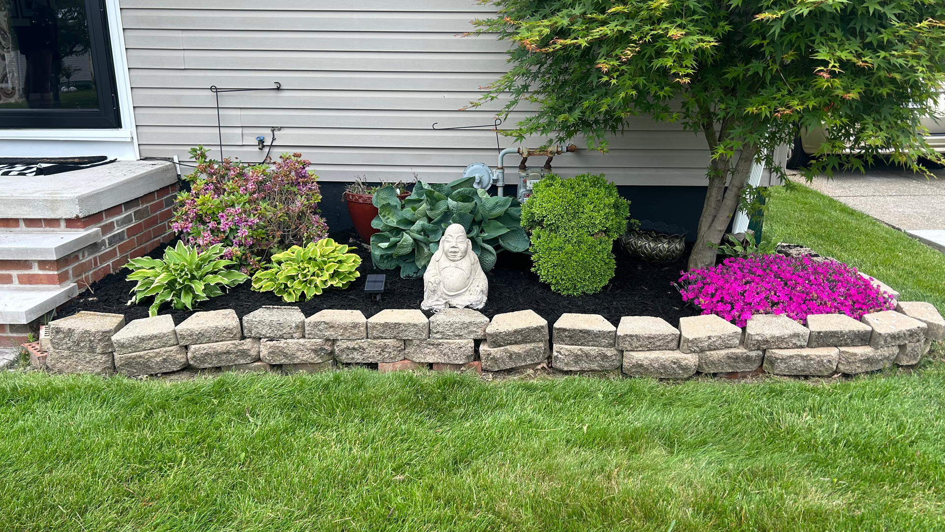 Stone-bordered flower bed with various plants and a statue, against a light-colored house. Green grass in the foreground.