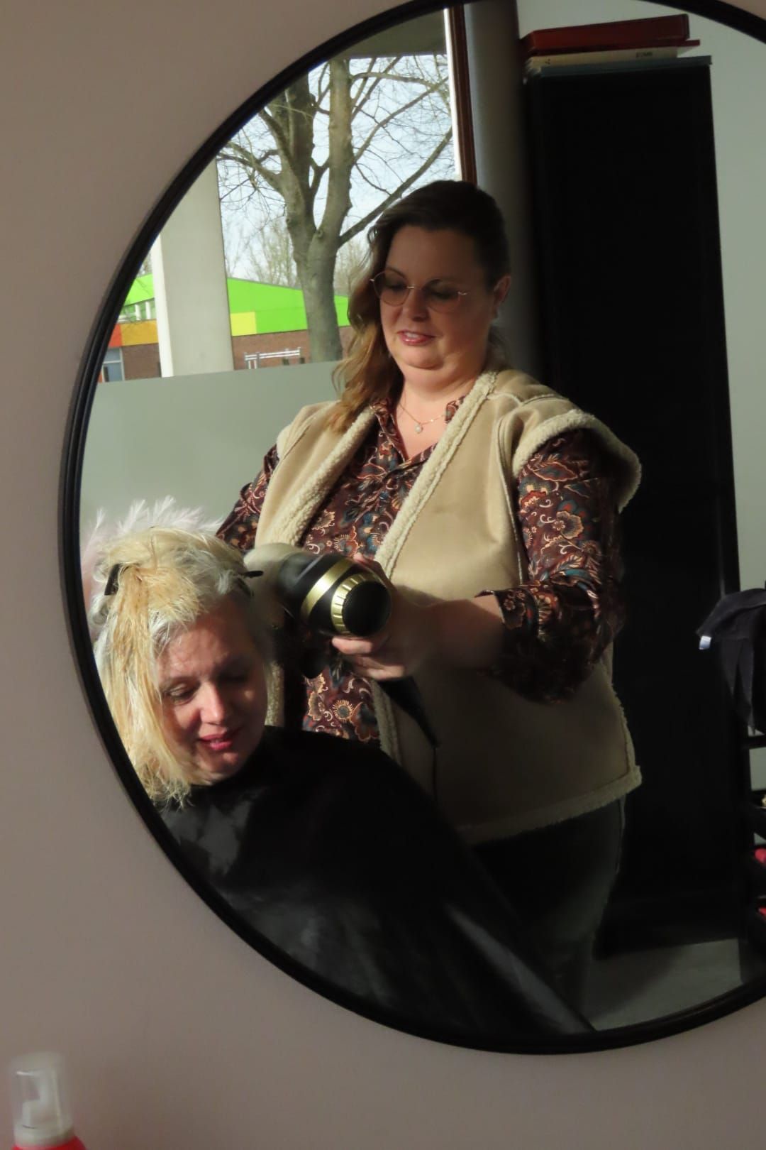 Een vrouw gebruikt een camera in een salonspiegel, met een zittende persoon weerspiegeld op de voorgrond.