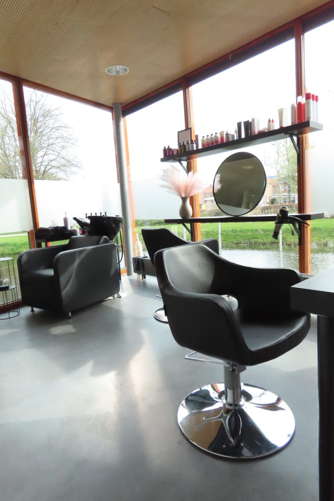 Modern interieur van een kapsalon met zwarte kappersstoelen, spiegels en grote ramen met uitzicht op groen.