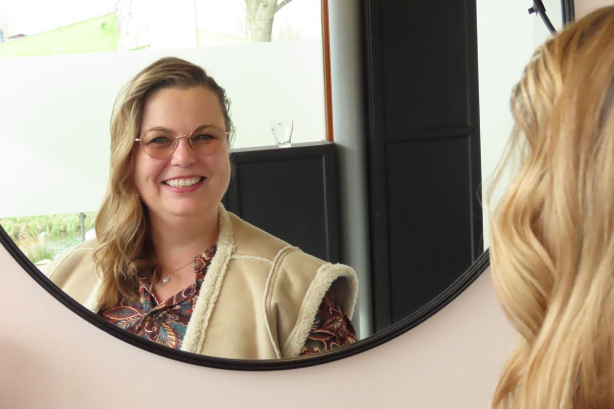 Vrouw lacht in een spiegel, draagt een bril en een beige trui in een lichte kamer.