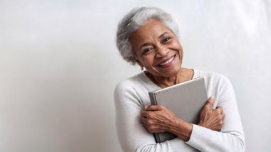 An elderly woman is hugging a book and smiling.