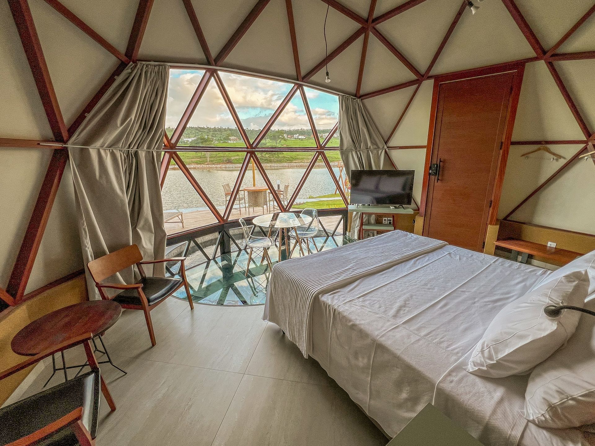 Quarto em forma de cúpula geodésica com cama branca, escrivaninha e grandes janelas com vista para uma paisagem verdejante.