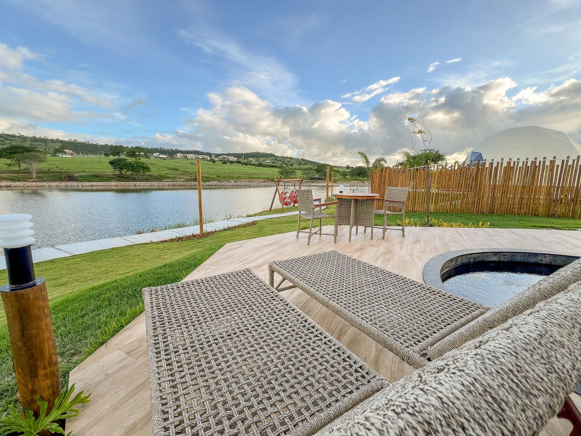 Pátio com vista para um lago e colinas, com cadeiras de vime, fogueira e céu nublado.