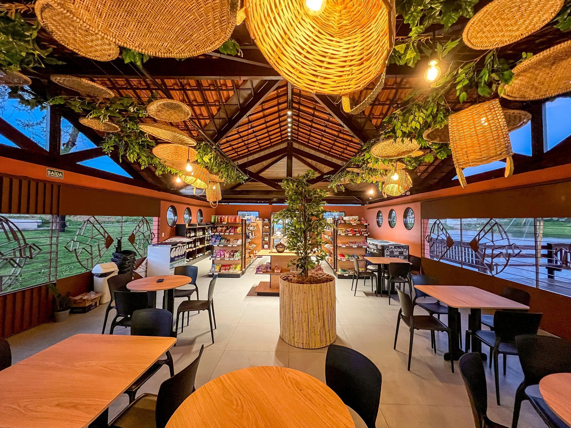 Restaurante ao ar livre com luminárias de teto trançadas, mesas de madeira e vista para o mar ao pôr do sol.