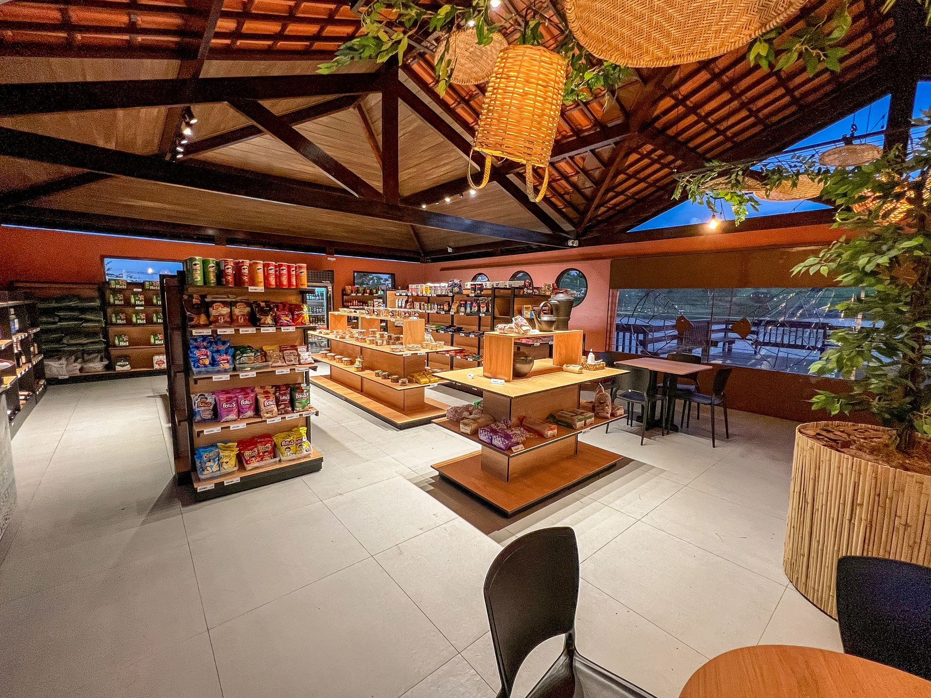 Café ao ar livre com vigas de madeira, luminárias de teto trançadas e mesas centrais com exposição de lanches e bebidas.