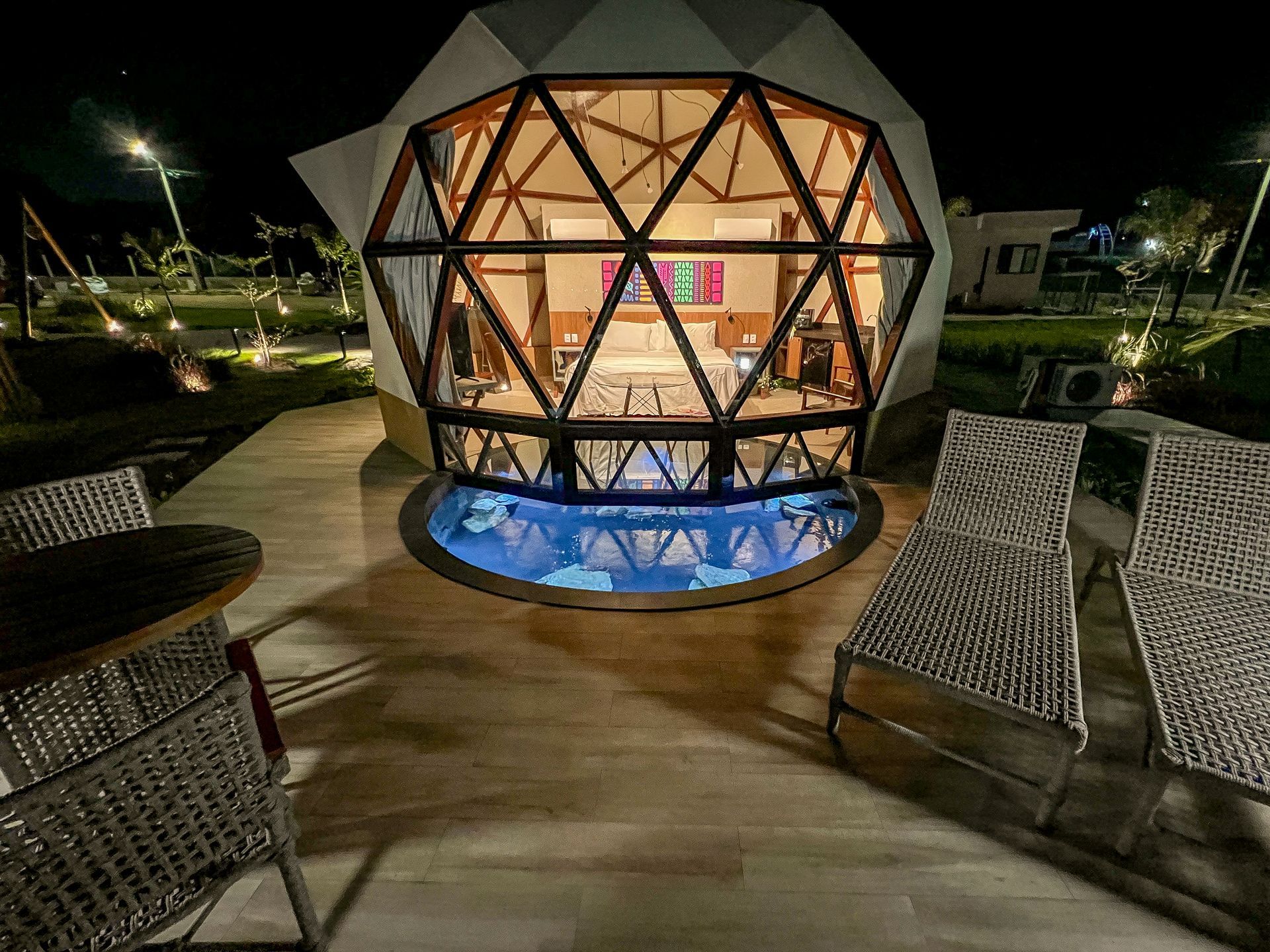 Pavilhão geodésico iluminado com luzes quentes e uma pequena piscina iluminada em azul à noite, visto de um pátio com cadeiras.
