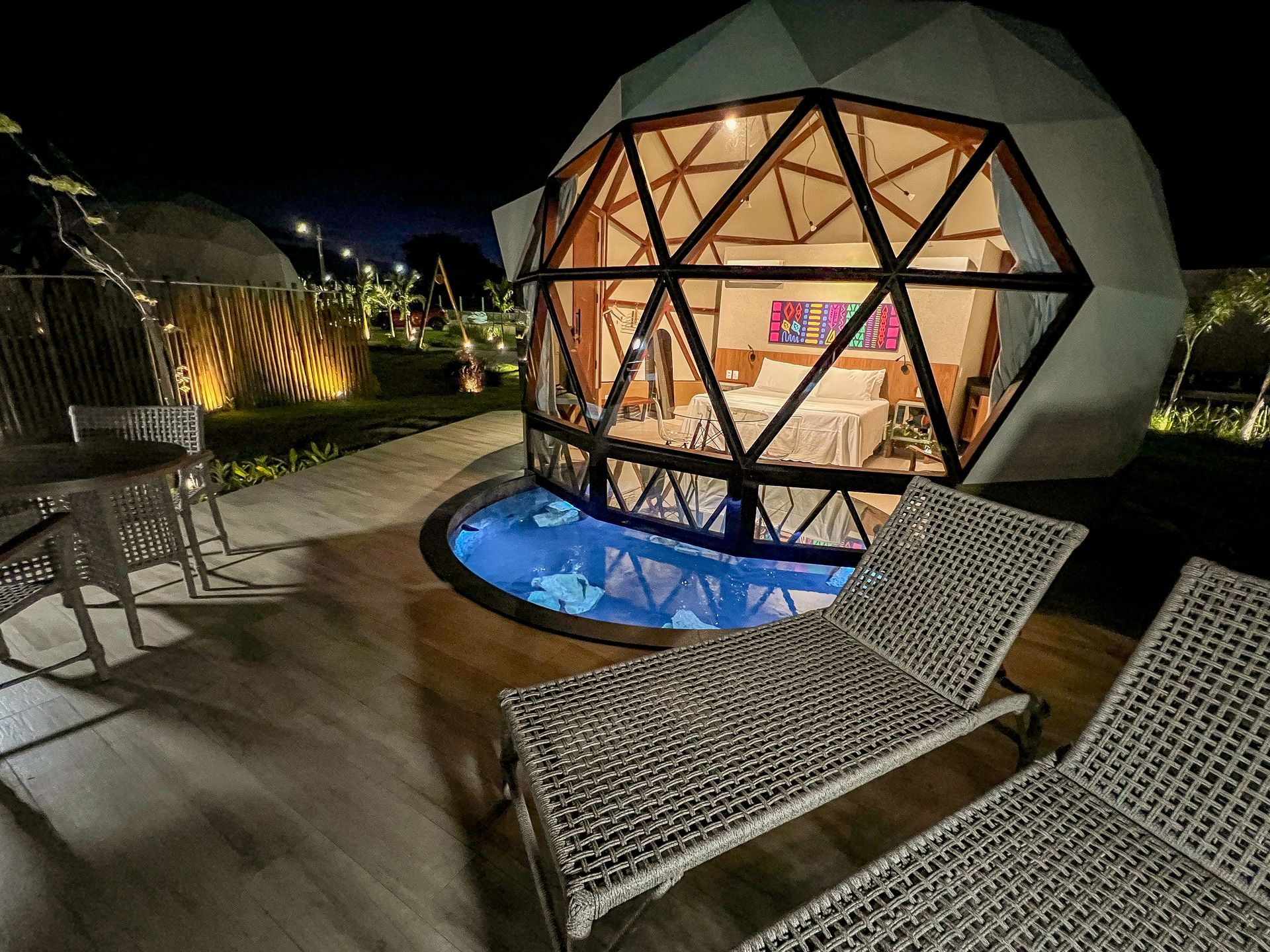 Pátio noturno com cúpula geodésica iluminada, banheira de hidromassagem brilhante e espreguiçadeiras de vime.