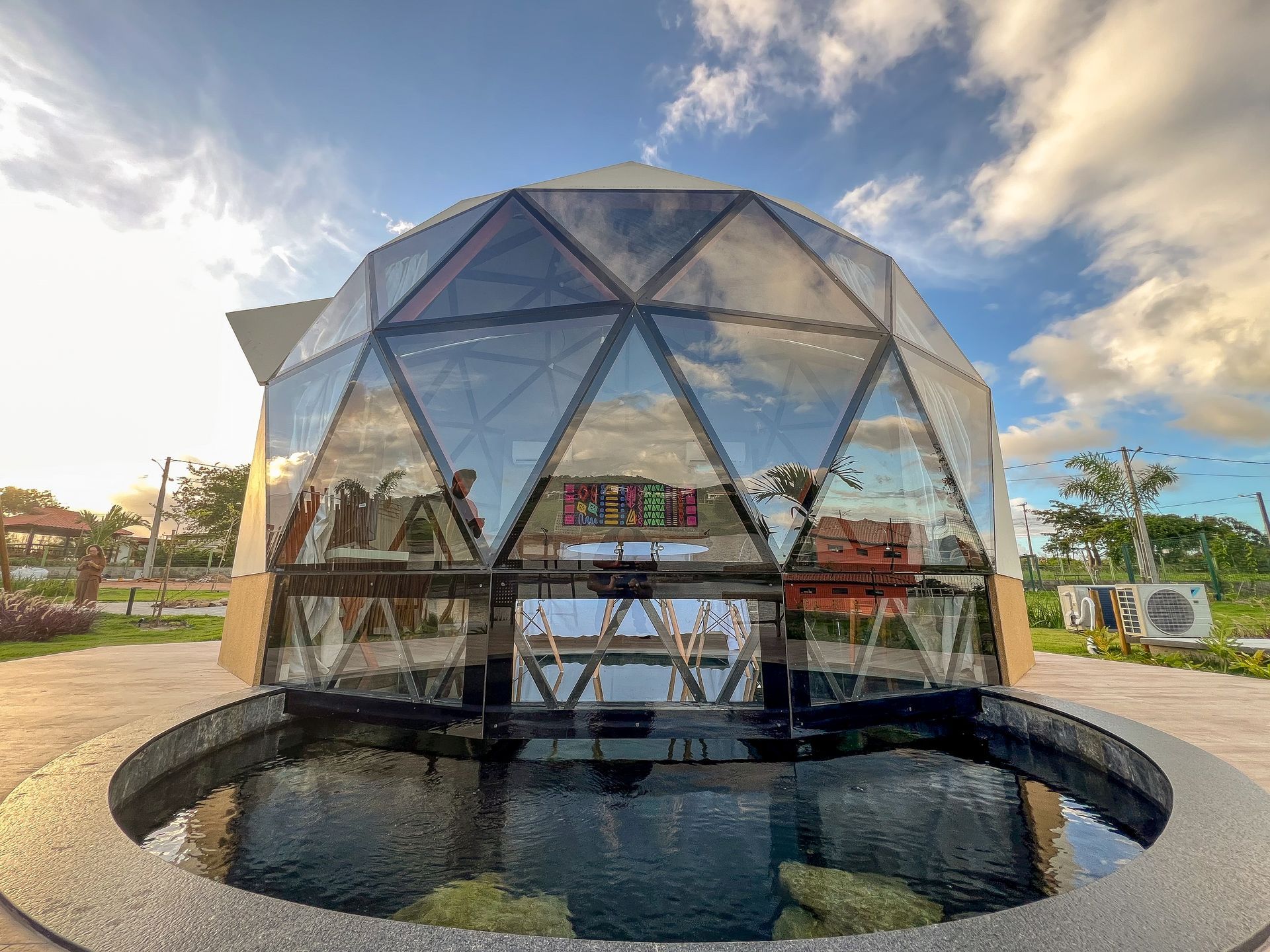Pavilhão geodésico de vidro sobre um espelho d'água circular sob um céu parcialmente nublado.