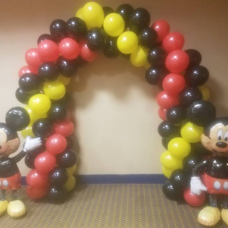 Two mickey mouse balloons are standing under a balloon arch