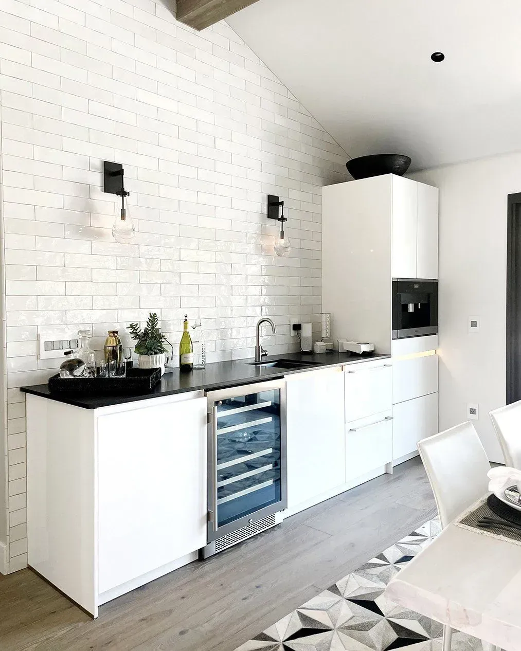 White kitchen with brick wall, black countertops, and wine fridge.