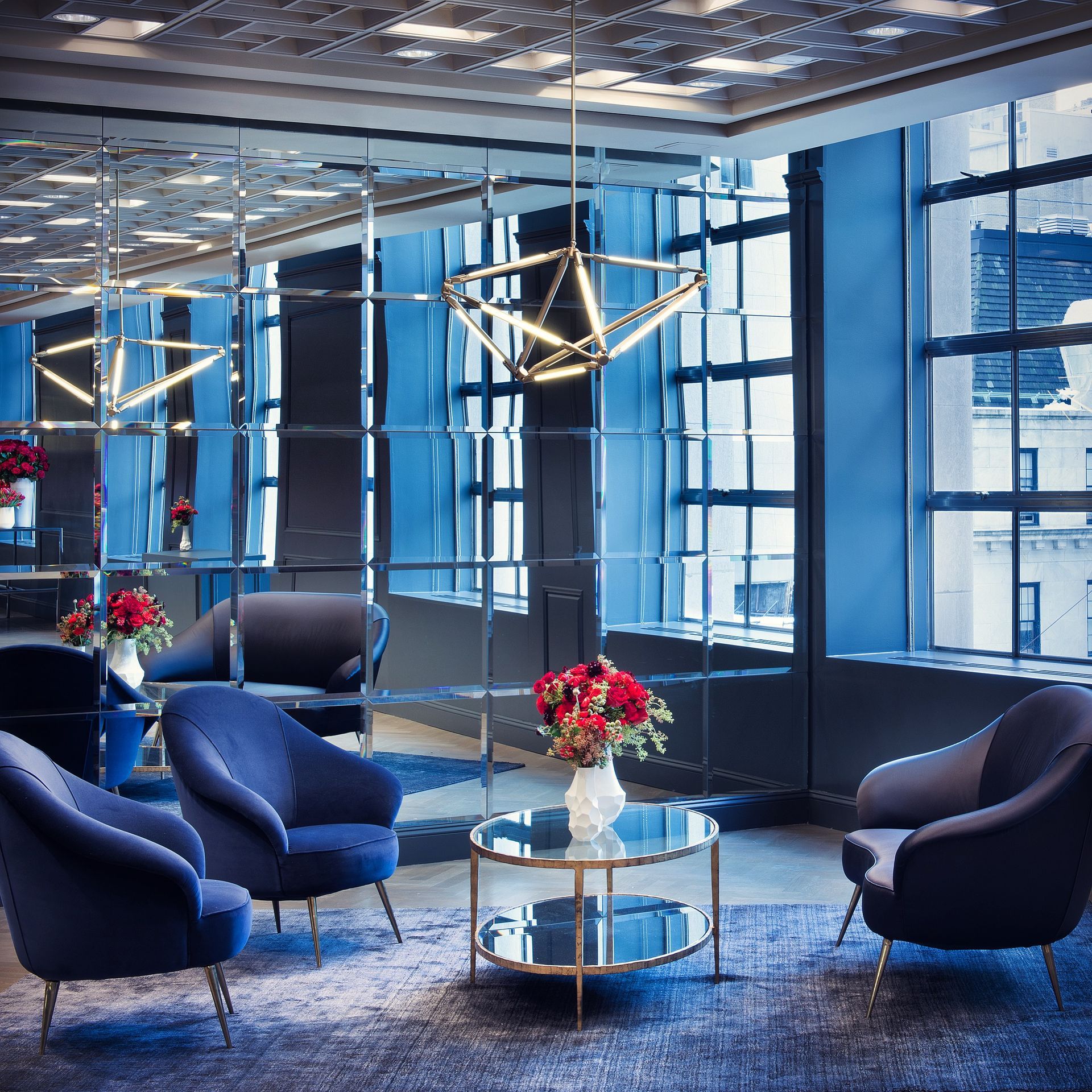 Modern lounge with blue chairs, glass tables, red flowers, and large windows in a stylish office lobby