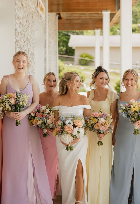 Five women in pastel dresses holding bouquets at a wedding venue, smiling on a porch