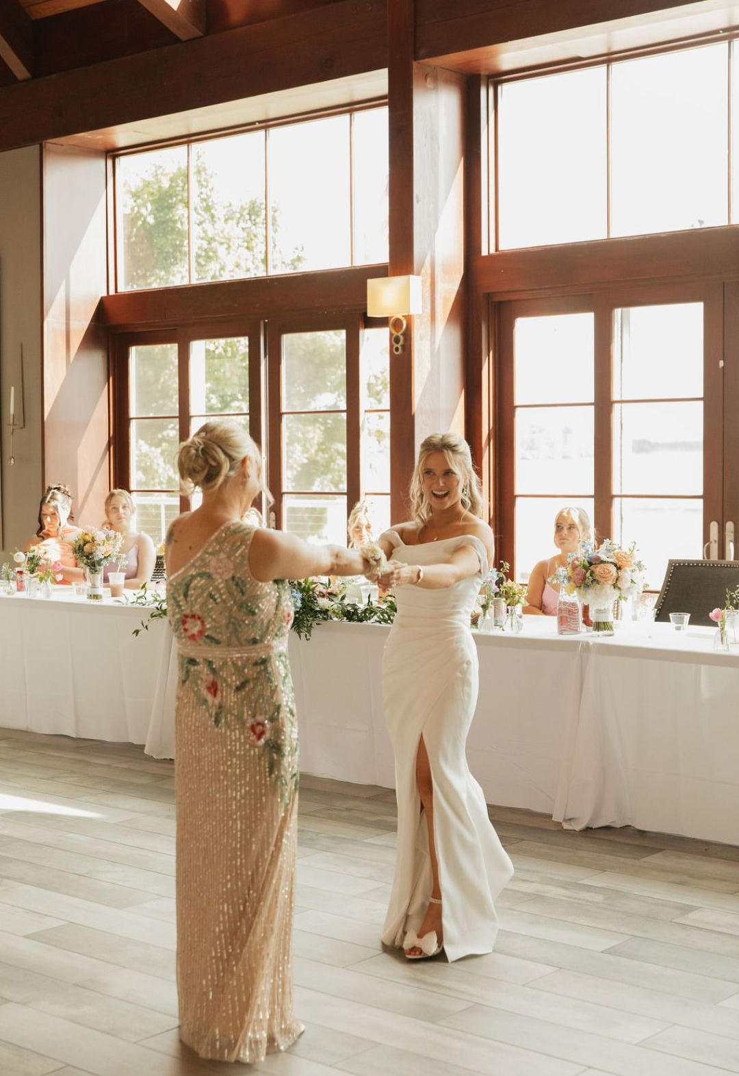 Two women in formal dresses dance at a bright wedding reception with floral tables and tall windows
