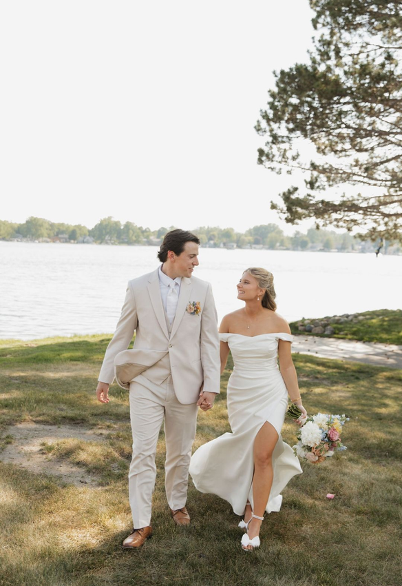 Couple in wedding attire walking together by a lakeside, smiling under trees.