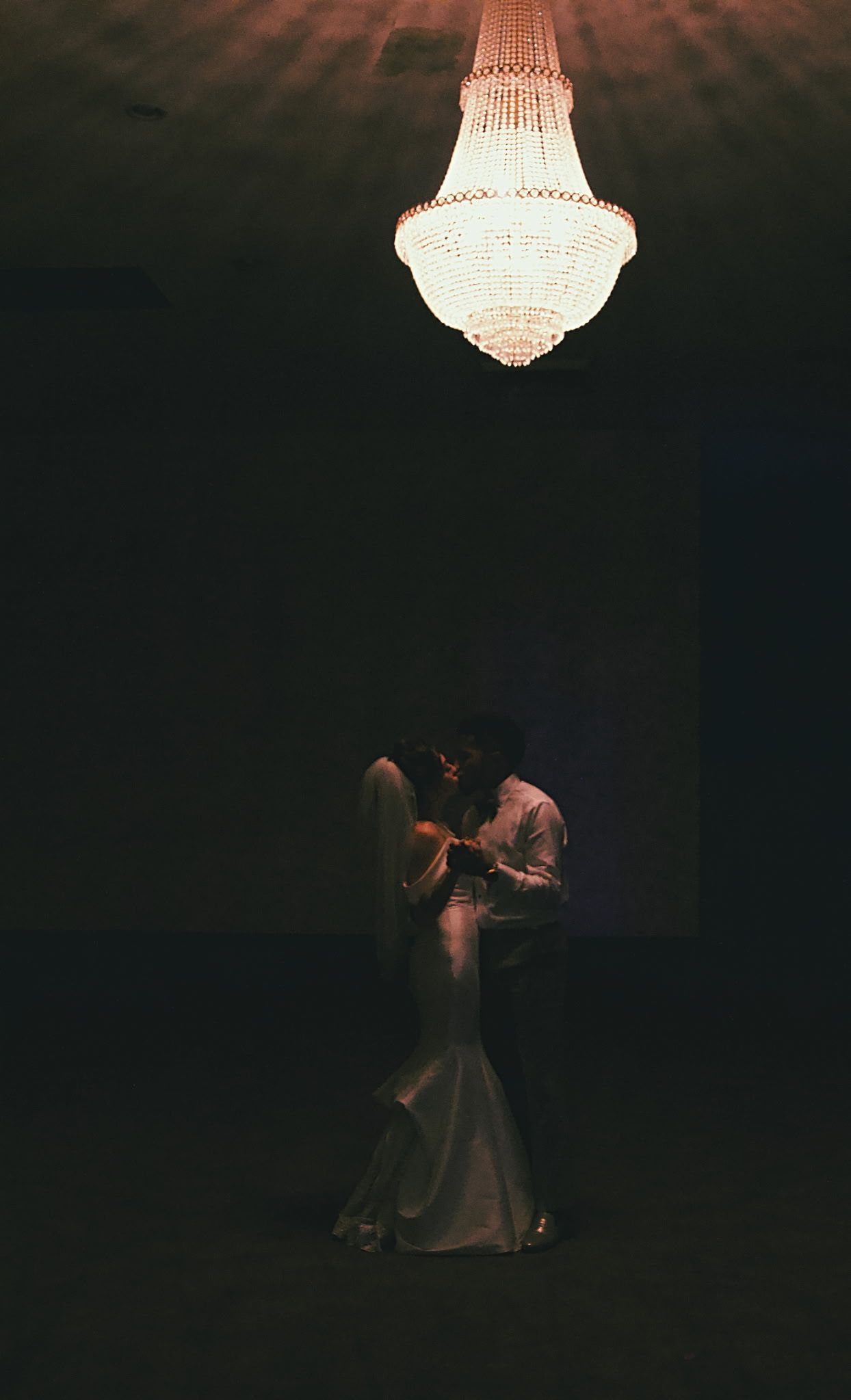 A couple dancing beneath a glowing chandelier in a dark ballroom.