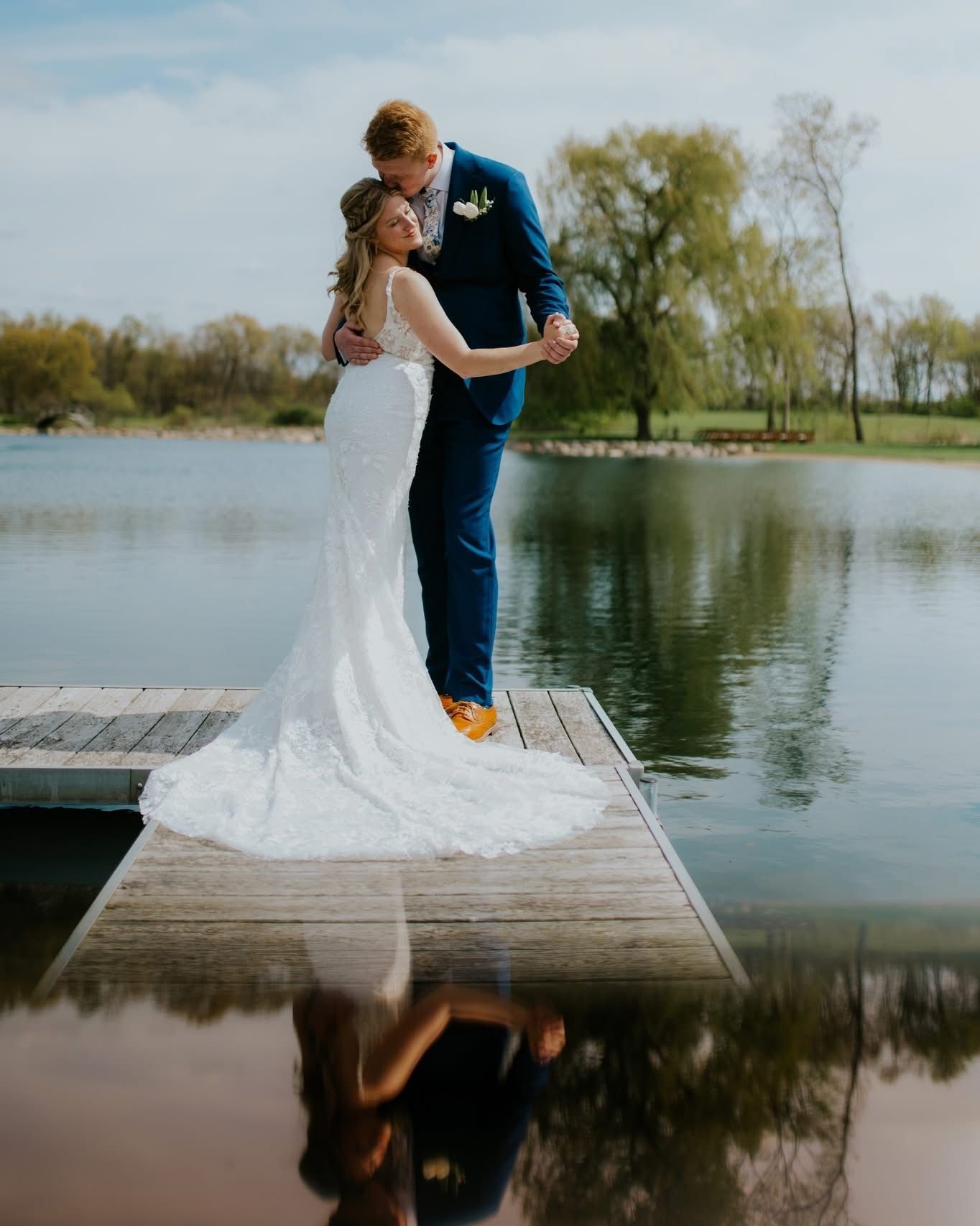 Couple embracing on a wooden dock by a lake, with a wedding dress and reflection in the water.