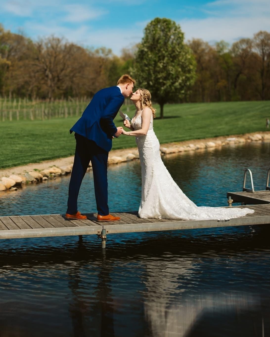 Bride and groom kissing on a dock by a pond, with trees and grass in the background.