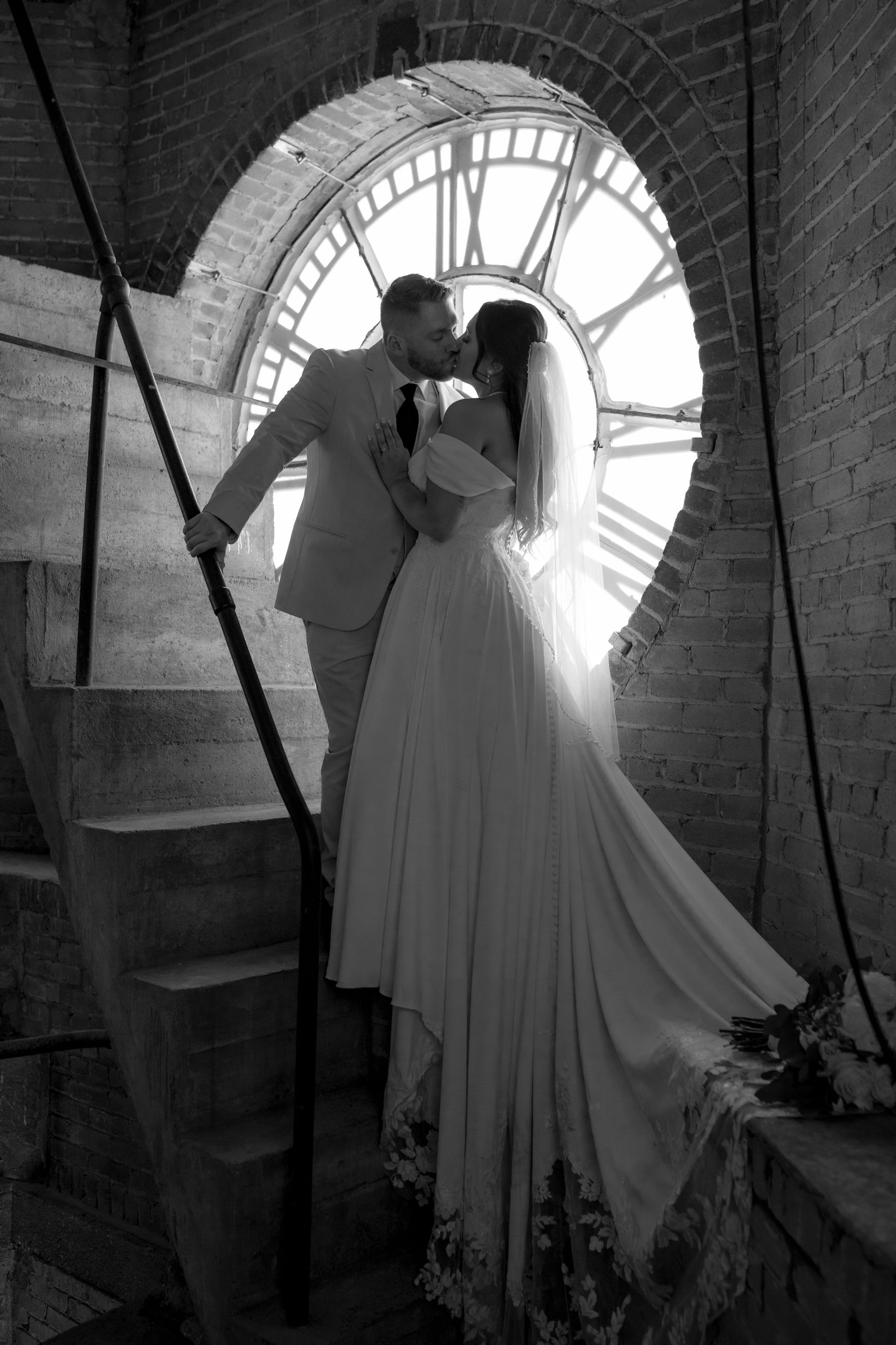 Couple embracing by a circular window in a brick stairwell, bride in flowing dress and groom in suit
