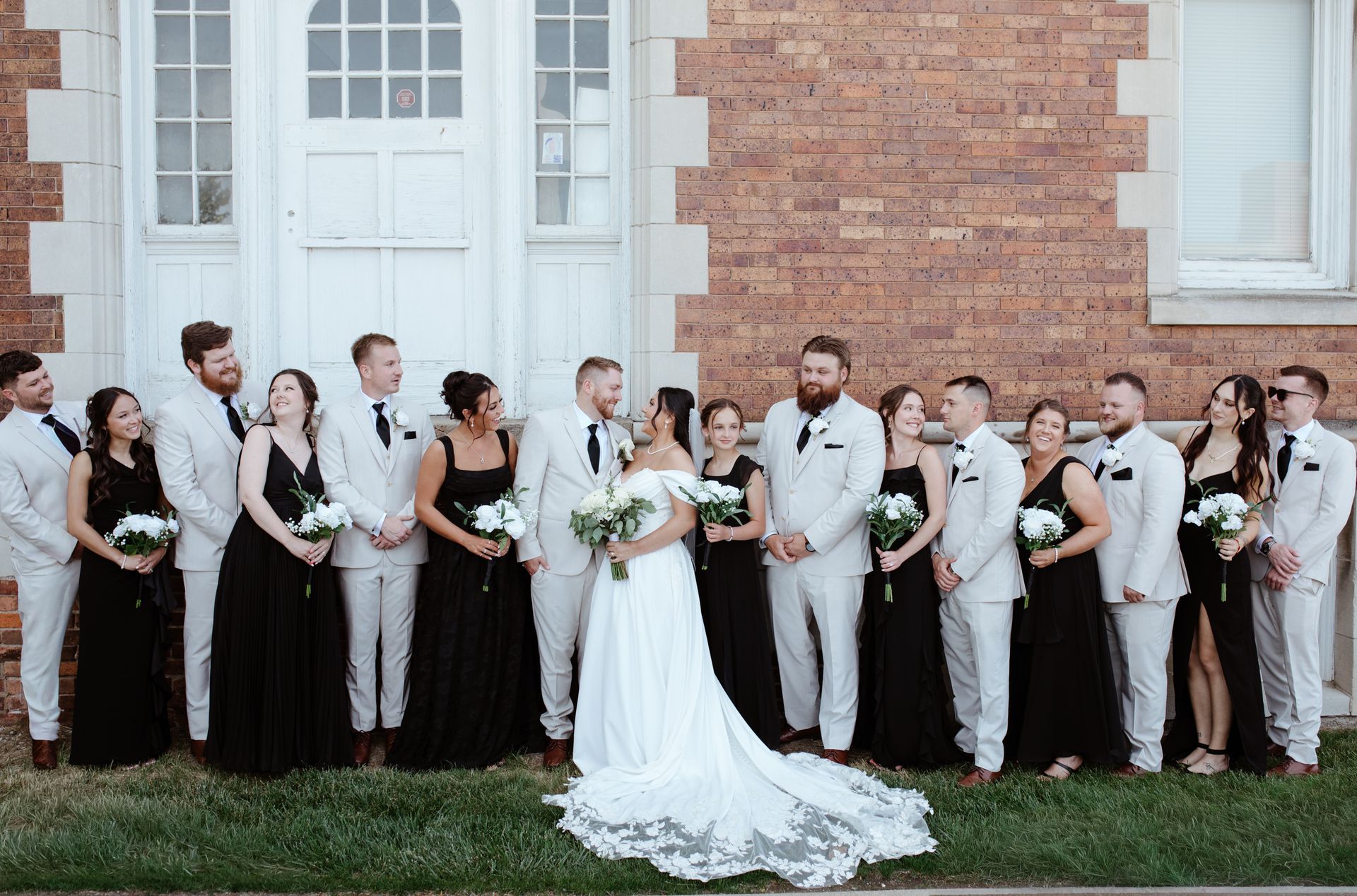 Wedding party posing in front of a brick building, with the bride in a white gown at center.