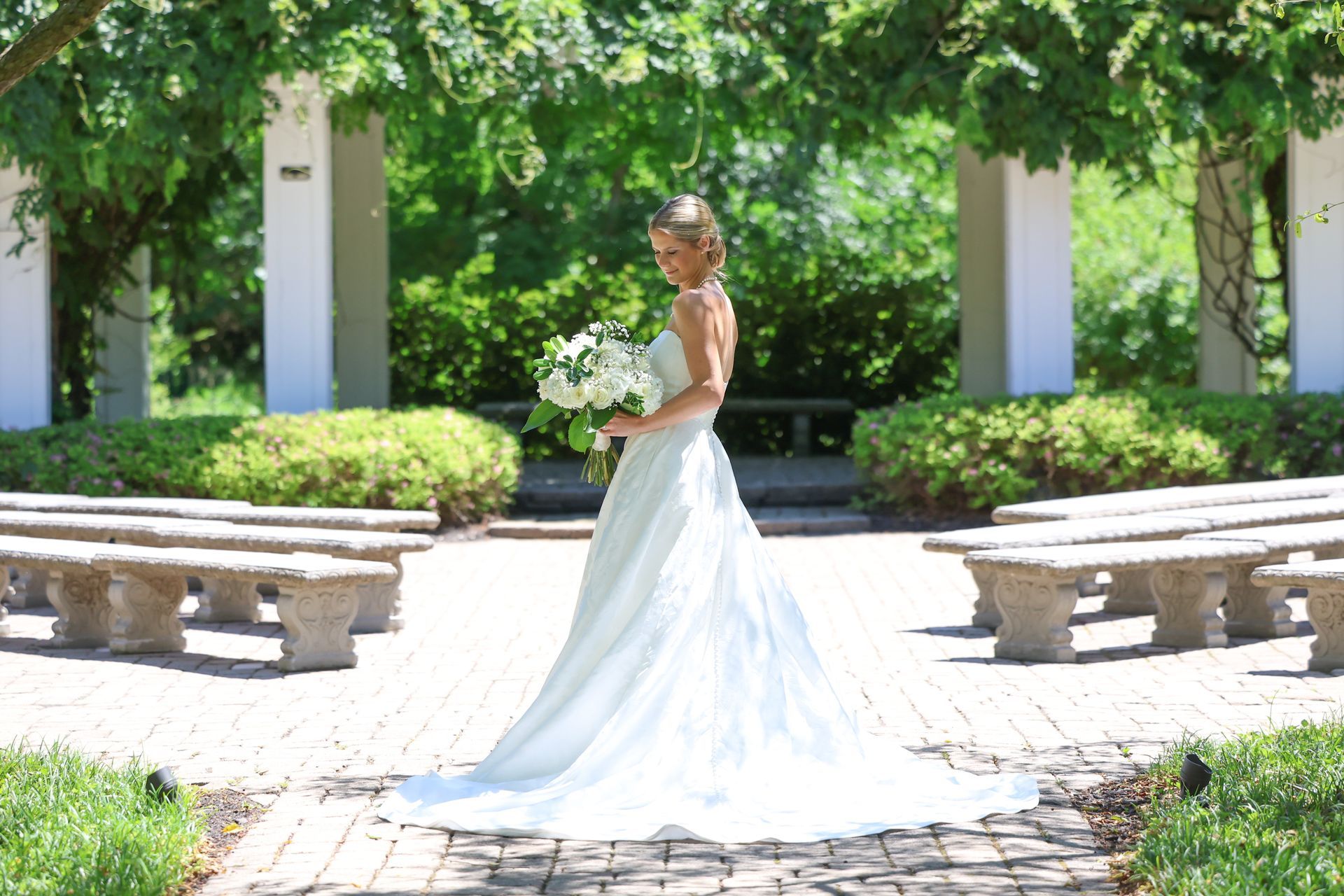 Bride in a white gown holding a bouquet in a sunlit garden walkway between benches