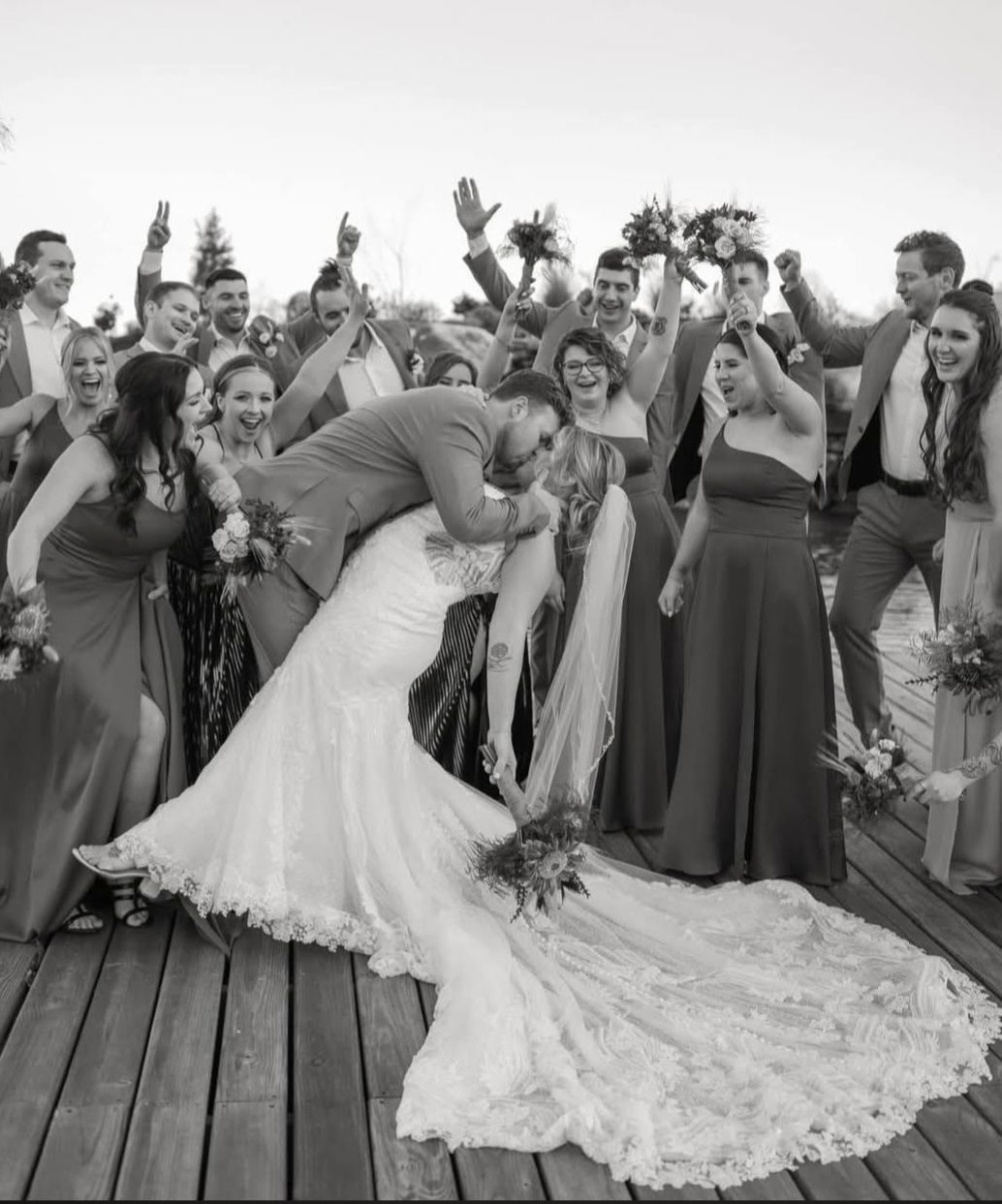 Bride and groom kiss as guests cheer on a wooden deck, the bride’s long train spread across the floor.