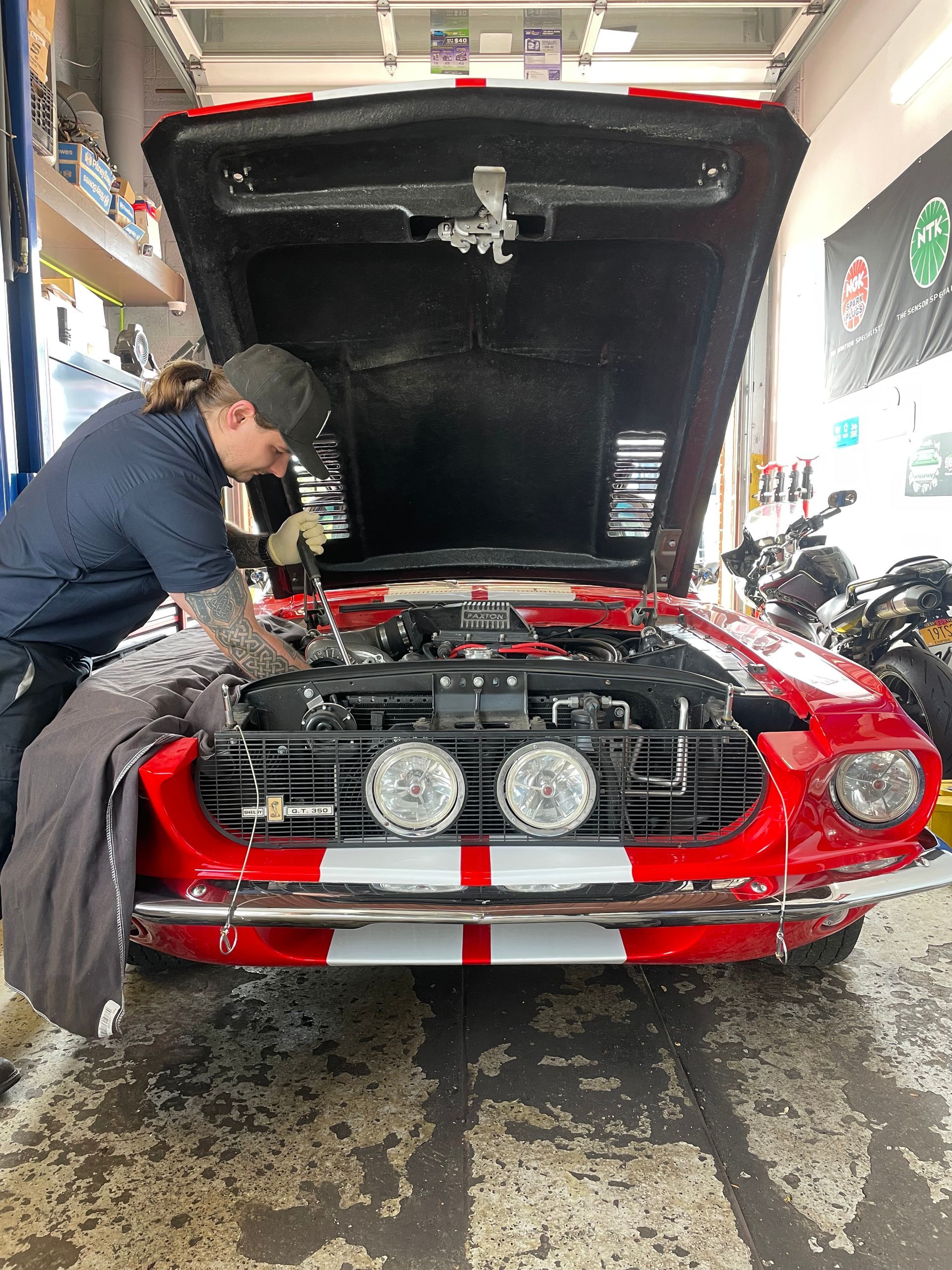 Mechanic working on the engine of a bright red vintage car with white stripes and round headlights in a garage.